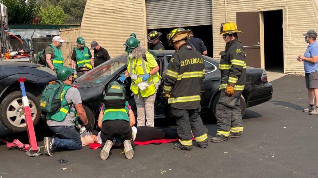 Three people wearing Community Emergency Response Team vests and two Cloverdale fireman demonstrate emergency first aid on a willing volunteer, who is lying on the pavement next to a car as if in an automobile accident.